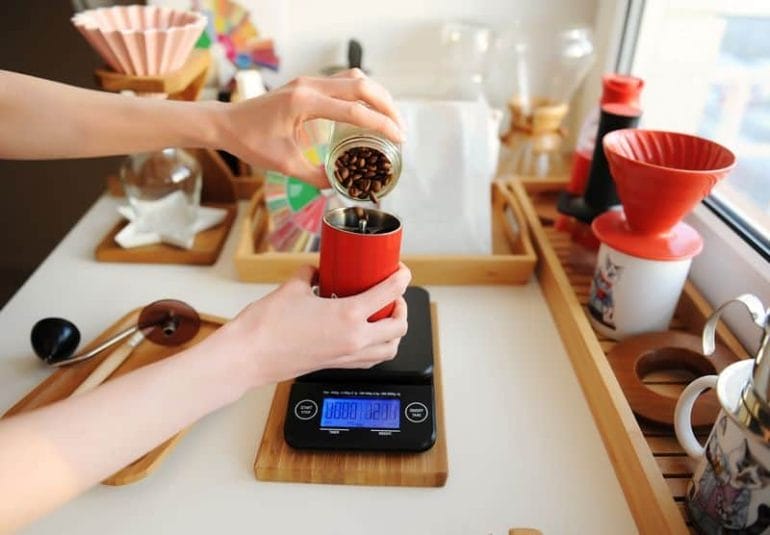 Pouring coffee beans into red manual coffee grinder on electronic scale