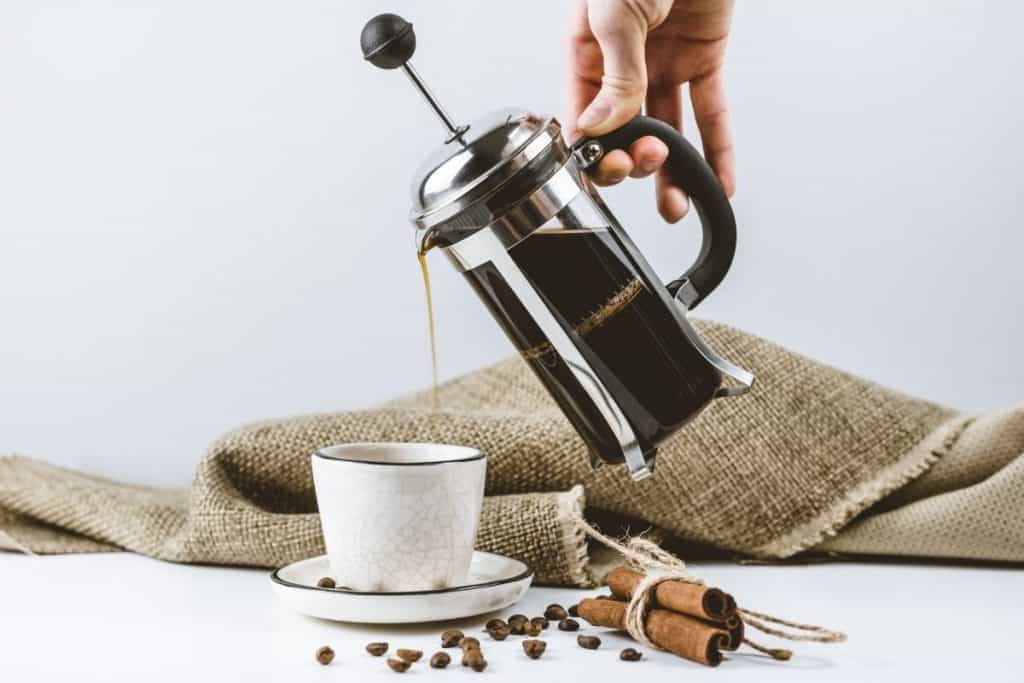 Glass french press coffee maker full of brewed coffee pouring coffee into white coffee cup and saucer.