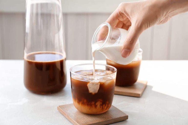Carafe of cold brew coffee and woman pouring milk into glass of cold brew