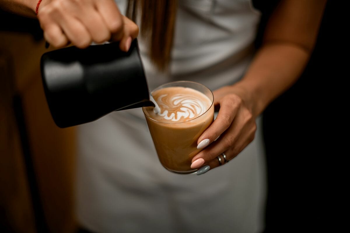 Close-up view of woman gently pouring milk from a latte milk jug into glass making latte art.