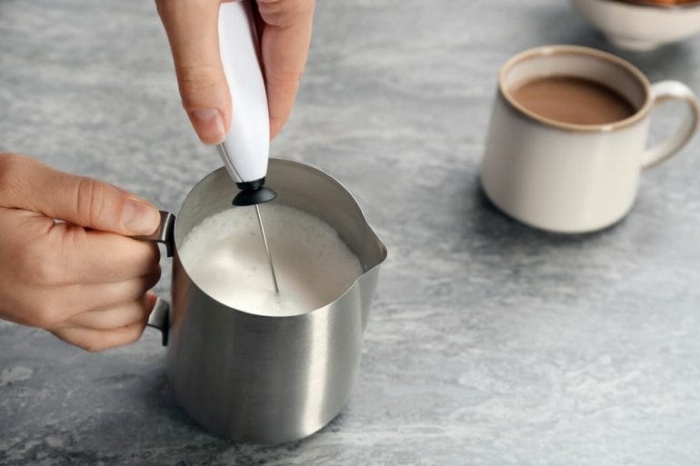 Woman using handheld milk frother in pitcher near cup of coffee on table.