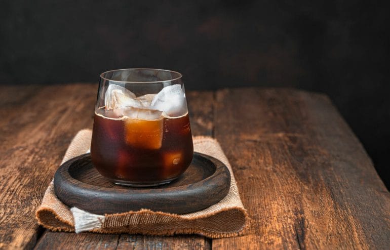 Black iced coffee in a clear, steamed glass on a brown background.