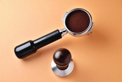 coffee portafilter with natural ground coffee and wooden coffee tamper on an orange background close-up.