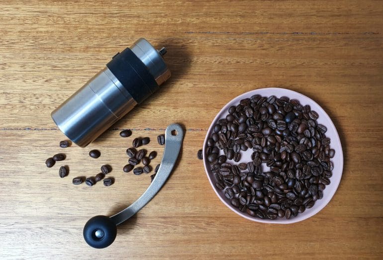 Stainless steel manual coffee grinder with bowl of roasted coffee beans.