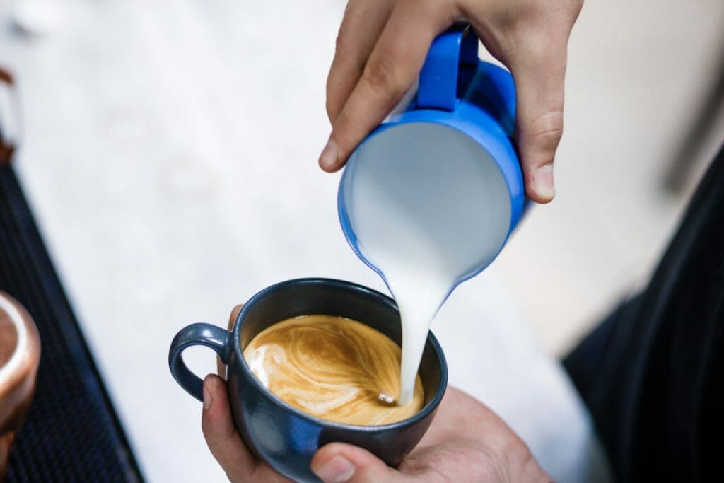 Person holding blue milk jug pouring into cup of coffee.