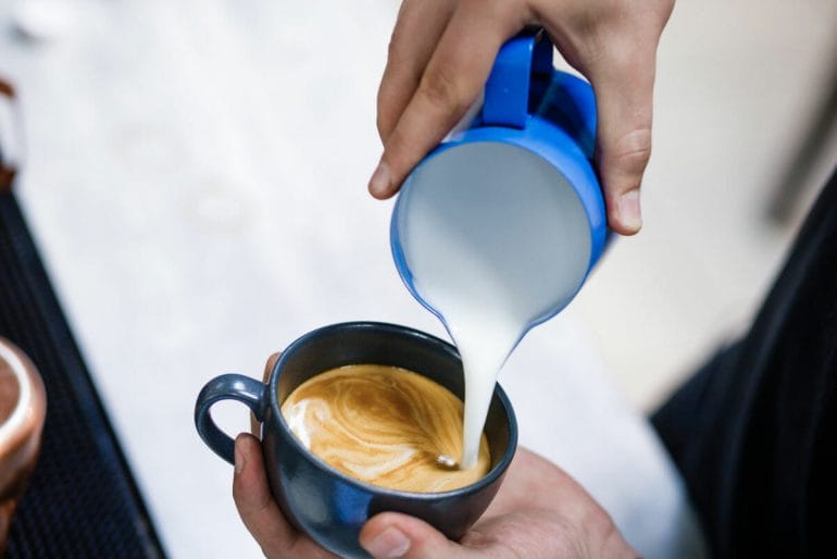 Person holding blue milk jug pouring into cup of coffee.