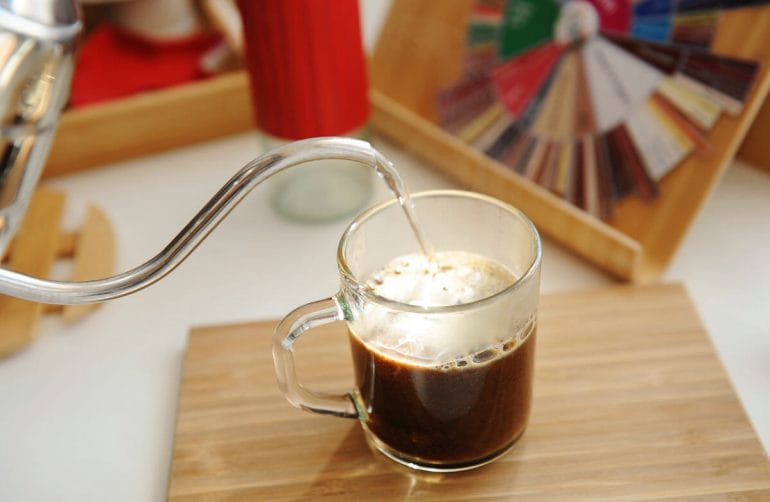 Americano coffee in mug. Pouring water from gooseneck kettle into a glassy cup of espresso coffee.