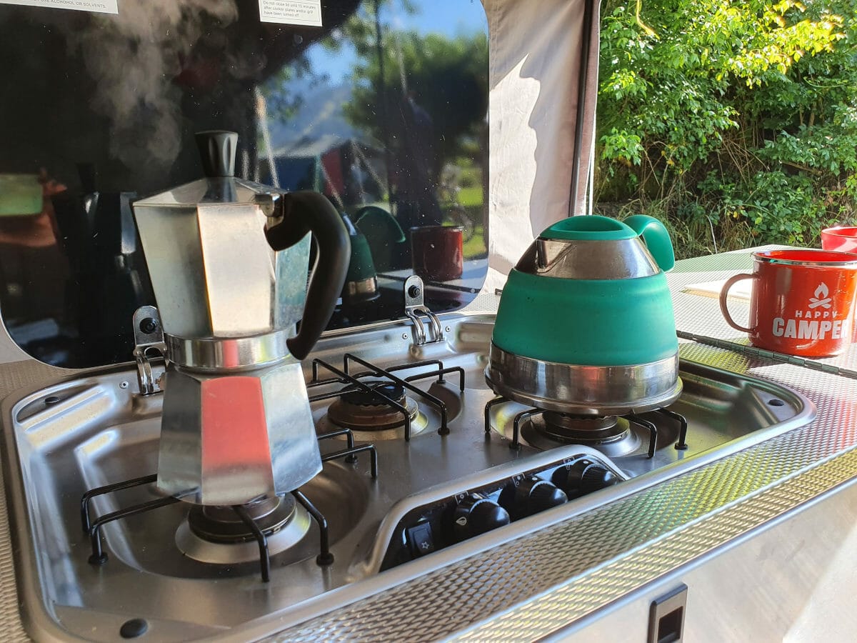 Bialetti moka pot on camp stove next to collapsible kettle and coffee mug.